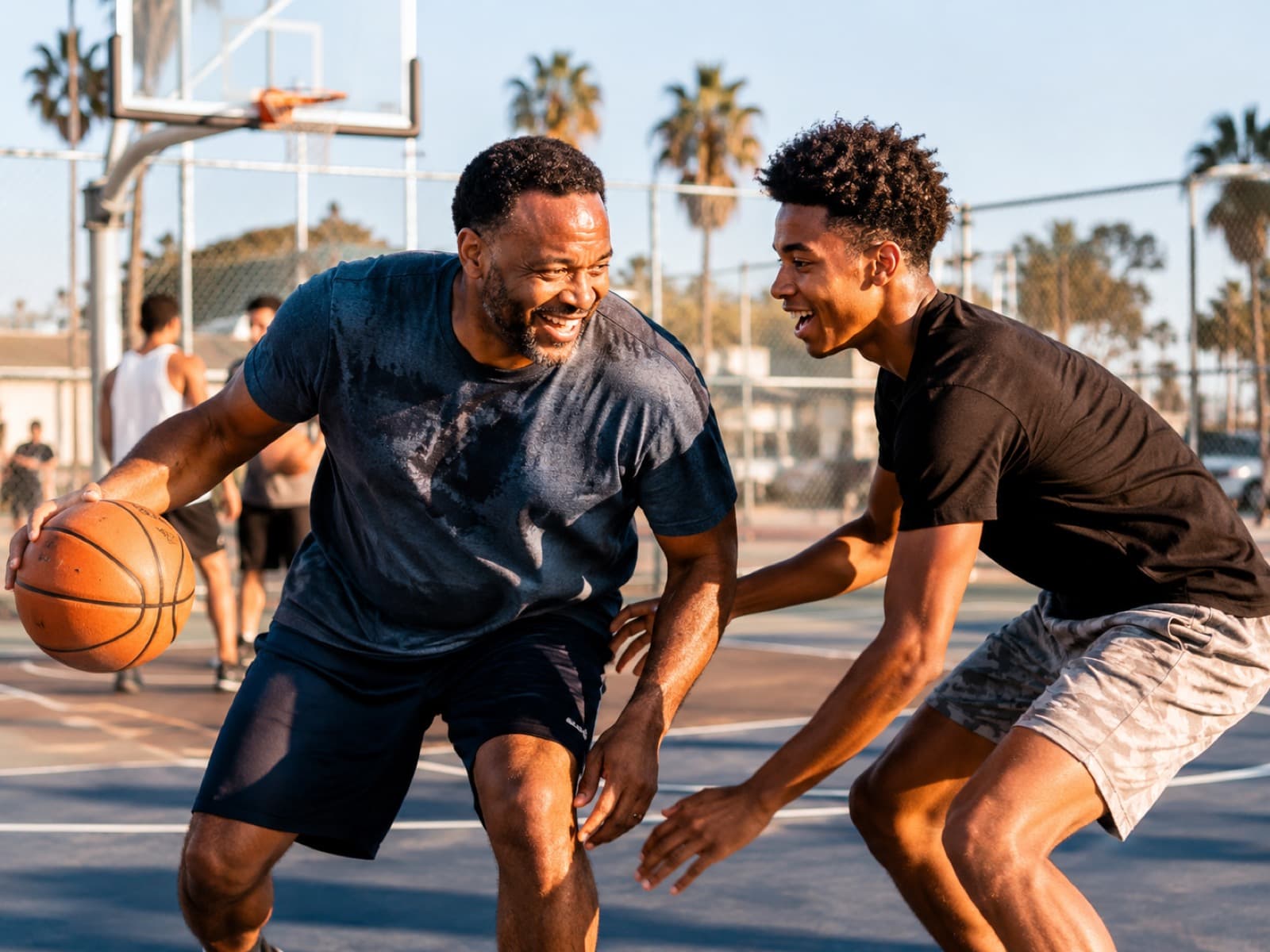 An older man and a teenager laughing during a friendly game of basketball on an outdoor court at golden hour.