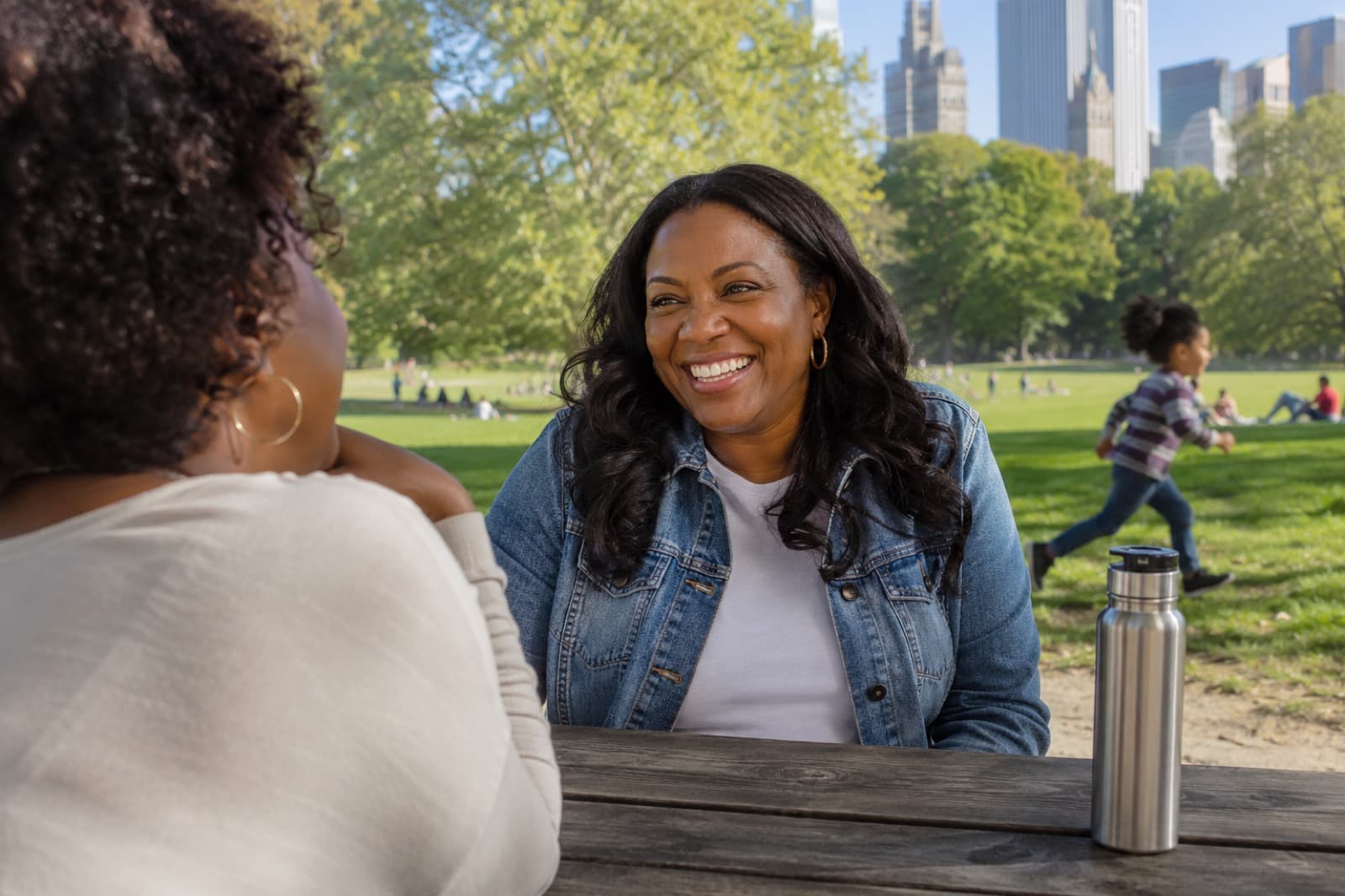 Two friends laughing together at a park table on a sunny afternoon, a city skyline behind them.